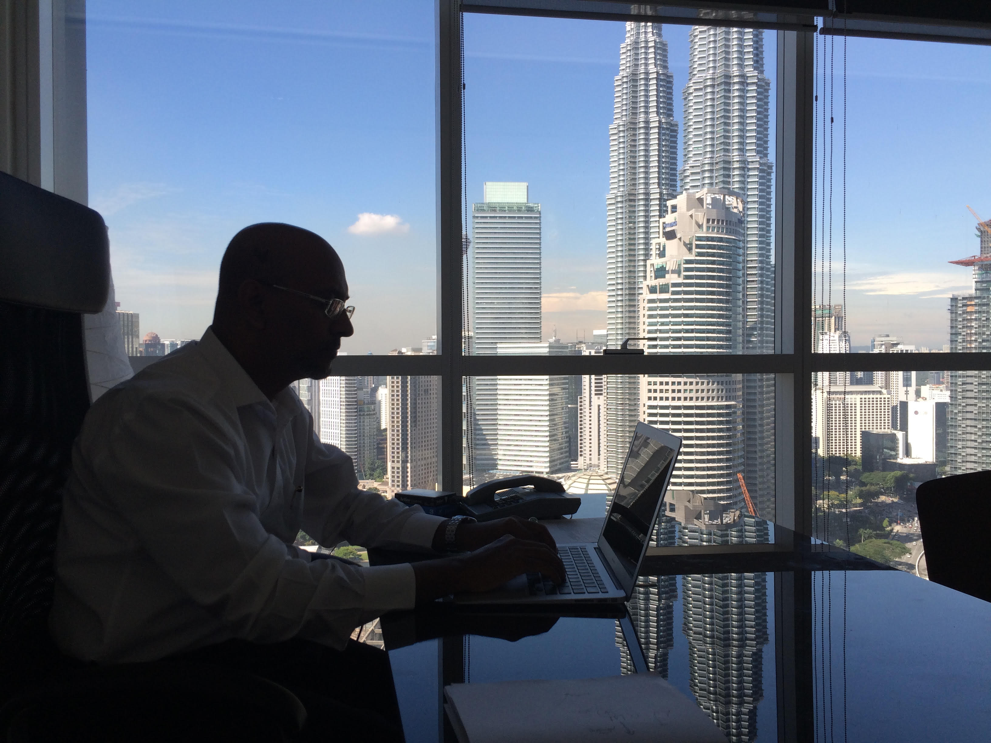 Ahamed Kameel Mydin Meera working at a laptop, the Kuala Lumpur skyline and the Petronas Towers through the window behind him